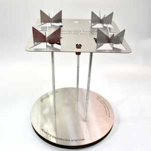 May include: A silver enameling station with four metal prongs and a round base. The base is labeled "Pearlys Enameling Station".