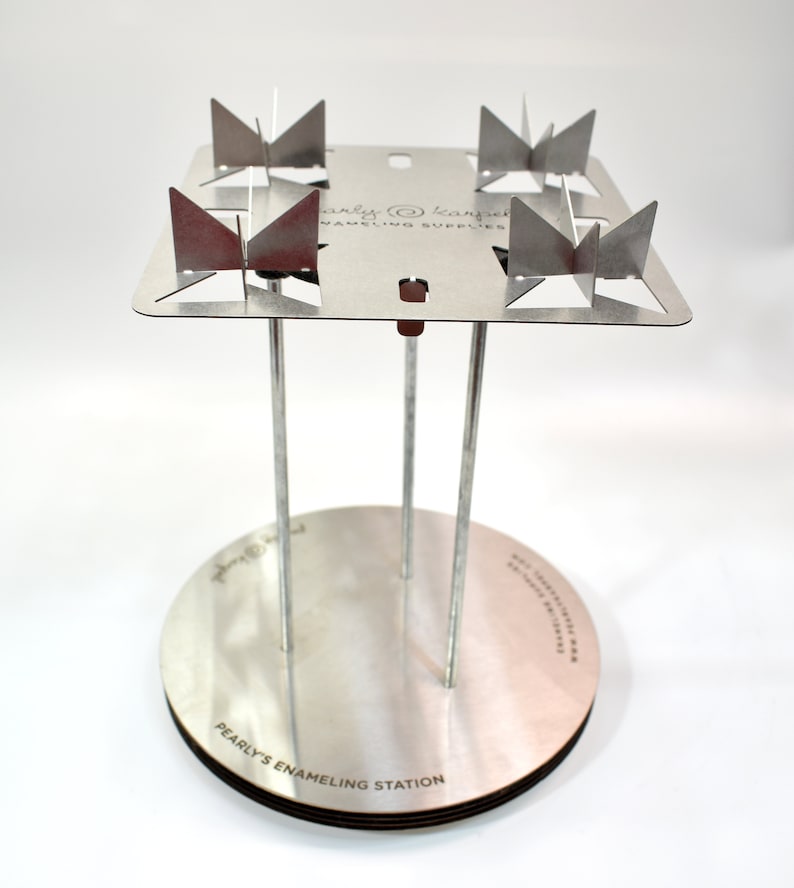 May include: A silver enameling station with four metal prongs and a round base. The base is labeled "Pearlys Enameling Station".