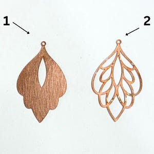 May include: Two copper-colored metal earring pendants. One is a solid teardrop shape with an oval cutout. The other is a filigree design with a similar teardrop shape, featuring an openwork pattern of leaves.
