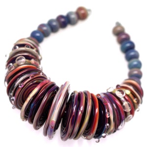 Op de afbeelding: Een kleurrijke glazen ketting met een spiraalvormig ontwerp. De ketting is gemaakt van kleine, ronde kralen in tinten rood, paars en blauw. De kralen zijn in een spiraalvorm gerangschikt, waardoor een uniek en opvallend ontwerp ontstaat.