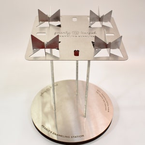 May include: A silver-colored enameling station with a square top and four metal supports. The top has four angled metal pieces. The base is round with the text "PEARLY'S ENAMELING STATION". The brand name "pearly @ karpel" is also visible.
