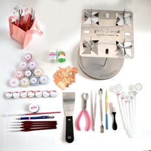 Puede incluir: Un conjunto de suministros para esmaltar que incluye un soporte de metal con cuatro molinetes, una variedad de polvos de esmalte de colores, un juego de pinceles, una espátula, pinzas, una pequeña herramienta de metal, un juego de cucharas de vidrio y dos botellas de líquido.