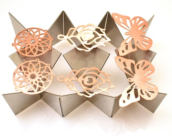 May include: Six copper-colored metal charms, including two mandala-like designs, two butterfly shapes, and two stylized feather designs, displayed on a gray triangular stand.