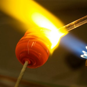 May include: A glassblowing torch with a blue flame heats a red glass bead on a metal rod.