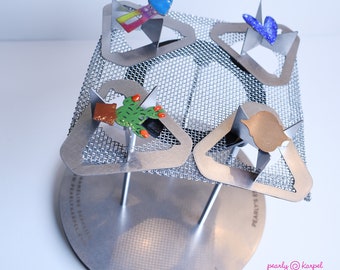 May include: A metal enameling rack with four triangular slots. Each slot holds a small enamel piece, including a cactus, a house, a butterfly, and a gold-colored piece. The rack is on a round base with the text "pearly@karpel enameling supplies" printed on it.