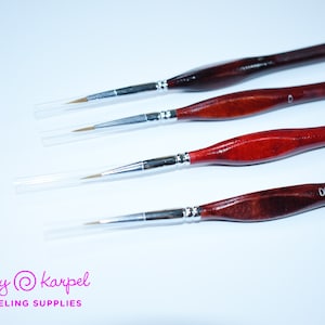 Puede incluir: Cuatro pinceles con mangos de madera y viroles de metal plateado. Los pinceles están etiquetados con 0, 00, 000 y 0000. Los pinceles están sobre una superficie blanca.