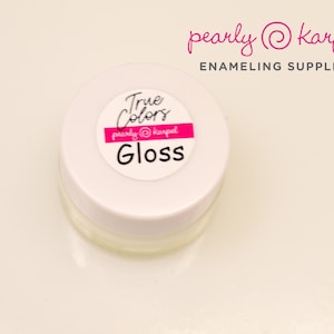 Puede incluir: Un pequeño frasco blanco con tapa transparente que contiene un esmalte blanco brillante. El frasco tiene una etiqueta rosa que dice "True Colors pearly @ karpel Gloss".