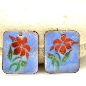 May include: Two square-shaped enamel charms with a blue background and a red flower with green leaves design.
