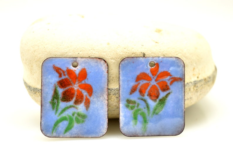 May include: Two square-shaped enamel charms with a blue background and a red flower with green leaves design.