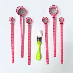 May include: Seven pink plastic tools with a mesh strainer at the top and a long handle. The tools are arranged in a row with a green brush in the center.