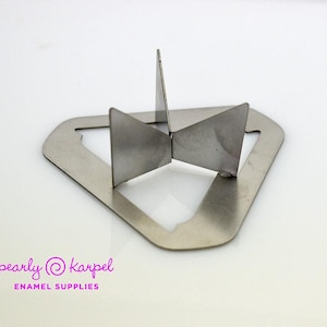 Peut inclure: Un support en émail métallique argenté avec un design géométrique. Le support a une base triangulaire avec des sections découpées et quatre supports inclinés. Le texte "pearly karpel ENAMEL SUPPLIES" est imprimé en rose et blanc.