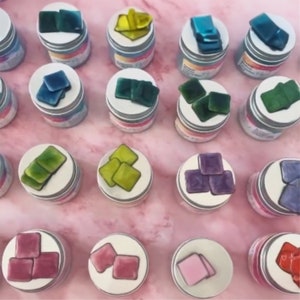 以下が含まれることがあります： 緑、青、紫、ピンク、黄など、さまざまな色の小さなガラスタイルの詰め合わせ。小さな丸い容器に並べてあります。