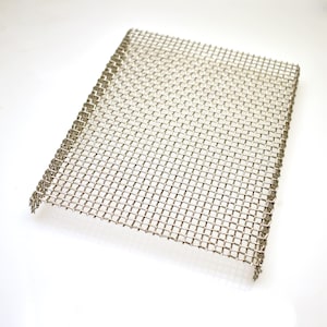May include: A rectangular piece of silver-colored metal wire mesh featuring a uniform grid pattern. The screen is constructed from thin, woven metallic strands that create a series of small, square-shaped openings across the entire surface area.