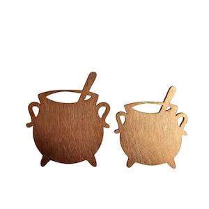 May include: Two gold-colored metal cauldron silhouettes with a spoon detail. The cauldrons are cut out and have a textured surface.
