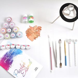 May include: An assortment of craft supplies, including small jars of colorful powders, bottles of liquid, and various tools. A color catalog with the text "Pearly's Enamel" is also present. The items are arranged on a white surface.