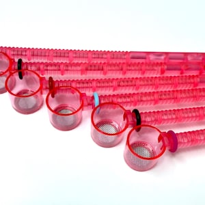 Peut inclure: Ensemble de bigoudis en plastique rose avec des coupelles transparentes et des filtres en maille. Les bigoudis sont disposés en diagonale, les coupelles face au spectateur. Chaque bigoudi a un anneau de couleur différente.