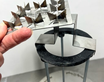 May include: A metal tool with a grid of triangular points, designed for creating decorative patterns in clay or other materials. The tool is attached to a metal stand with a black base.