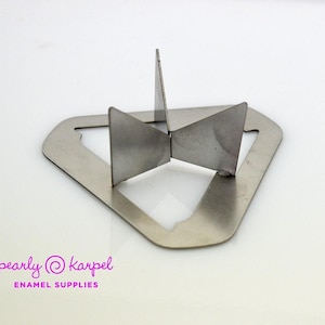 Könnte beinhalten: Ein silberfarbener Metalluntersetzer mit einem dreieckigen Basisrahmen und drei aufrechten, sich kreuzenden Metalllamellen, die aus der Mitte herausragen. Der Text "pearly karpel ENAMEL SUPPLIES" ist unten links in Pink aufgedruckt.
