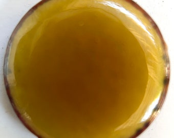 May include: A round, clear container filled with a golden yellow liquid. The container has a brown rim.
