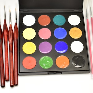 May include: A black palette with 16 round paint pots in various colors, including white, yellow, red, green, blue, and black. The palette is open and there are four paint brushes with wooden handles next to it.