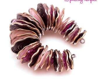 May include: A bracelet made of layered, scalloped glass beads in shades of pink, purple, and brown. The beads are arranged in a circular pattern and have a metallic finish.