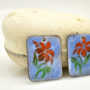 May include: Two rectangular enamel charms with a blue background and a white outline. Each charm features a red flower with green leaves.
