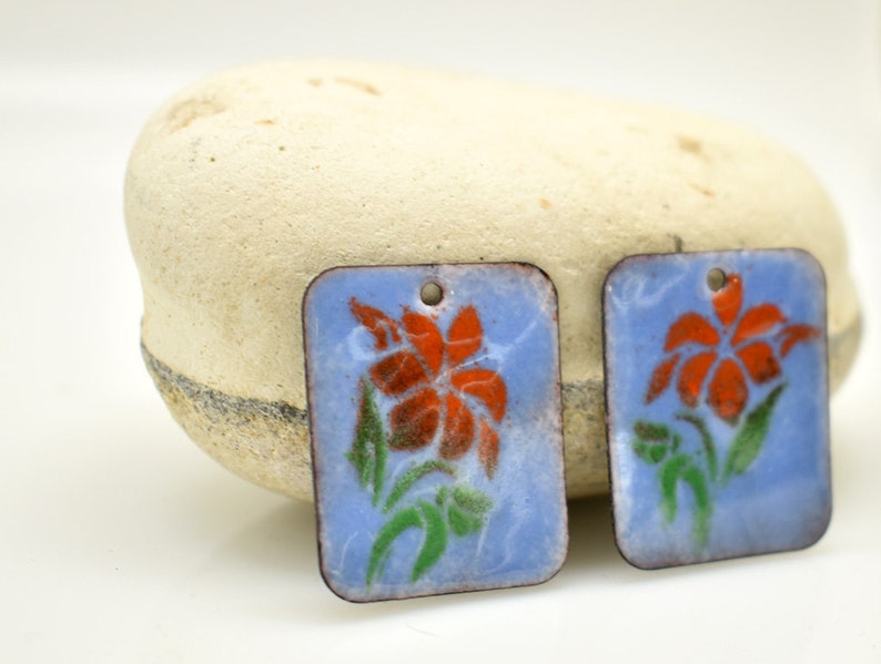 May include: Two rectangular enamel charms with a blue background and a white outline. Each charm features a red flower with green leaves.
