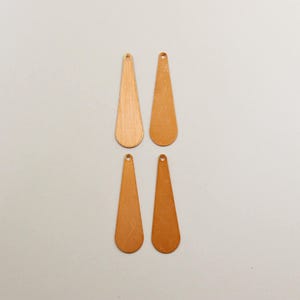 May include: Four elongated, teardrop-shaped copper pendants arranged in two rows. Each flat metal piece features a smooth, metallic finish and a single small circular hole at the narrow top end for attachment, showcasing a consistent warm tone.