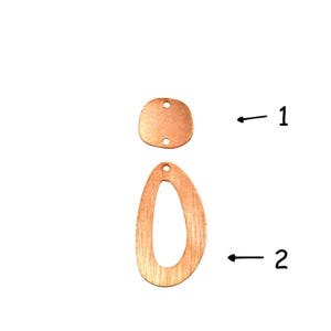 May include: Two copper-coloured metal jewellery components. Item 1 is a small, rounded square piece with two holes. Item 2 is a larger, elongated teardrop-shaped frame with a single hole at the top. Both pieces feature a brushed, textured finish.
