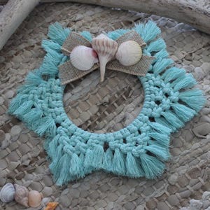 May include: A turquoise macrame wreath adorned with seashells and a burlap accent. The wreath is surrounded by various seashells and a piece of driftwood, creating a coastal theme. The background is a textured, neutral-toned mat.