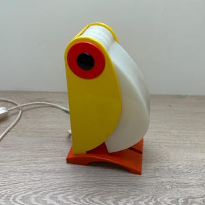 Peut inclure: Une lampe de table en forme de toucan avec un corps jaune vif, un bec blanc incurvé et un œil circulaire rouge avec un centre noir. La lampe repose sur une base orange et comprend un cordon d'alimentation blanc avec un interrupteur à l'arrière.