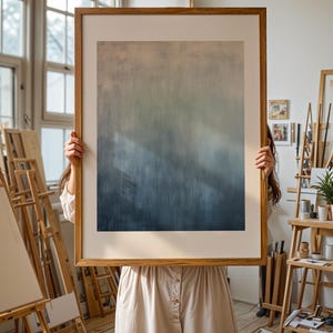 Pode incluir: Uma grande impress&atilde;o de arte abstrata emoldurada com um gradiente de cores do bege claro ao azul profundo. A obra de arte &eacute; segurada em frente a uma pessoa com um vestido de cor clara, em um ambiente de est&uacute;dio de artista. A moldura &eacute; de madeira clara.