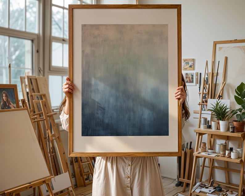 Pode incluir: Uma grande impress&atilde;o de arte abstrata emoldurada com um gradiente de cores do bege claro ao azul profundo. A obra de arte &eacute; segurada em frente a uma pessoa com um vestido de cor clara, em um ambiente de est&uacute;dio de artista. A moldura &eacute; de madeira clara.