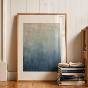 Pode incluir: Uma impress&atilde;o de arte abstrata emoldurada com um gradiente de tons azuis, verdes e bege. A obra de arte &eacute; exibida contra um fundo branco e repousa sobre um ch&atilde;o de madeira. Uma pilha de livros e uma cadeira de madeira tamb&eacute;m est&atilde;o na imagem.