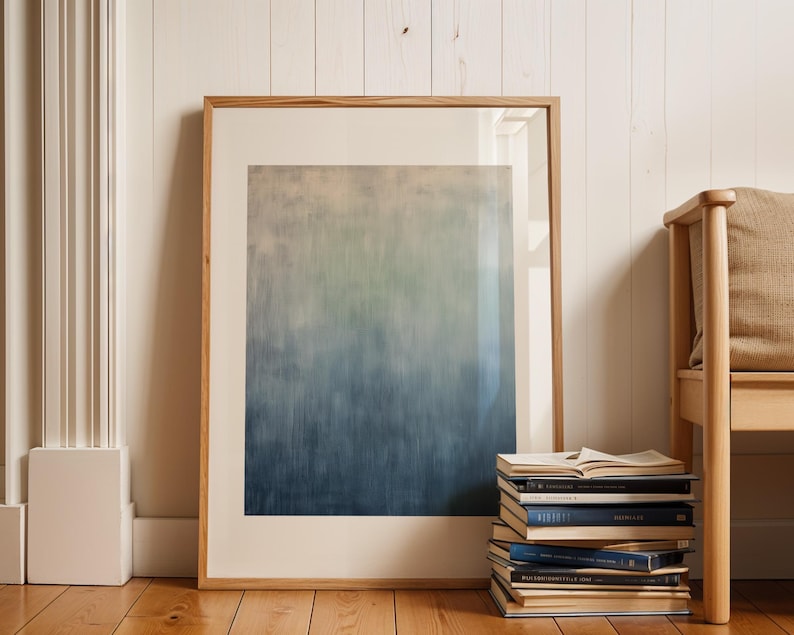 Pode incluir: Uma impress&atilde;o de arte abstrata emoldurada com um gradiente de tons azuis, verdes e bege. A obra de arte &eacute; exibida contra um fundo branco e repousa sobre um ch&atilde;o de madeira. Uma pilha de livros e uma cadeira de madeira tamb&eacute;m est&atilde;o na imagem.