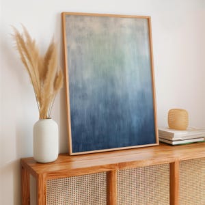 Pode incluir: Uma impress&atilde;o de arte abstrata emoldurada com um gradiente de tons azuis e verdes. A obra de arte &eacute; exibida em um arm&aacute;rio de madeira, ao lado de um vaso branco com capim-dos-pampas seco e uma pilha de livros. A moldura &eacute; de madeira clara.