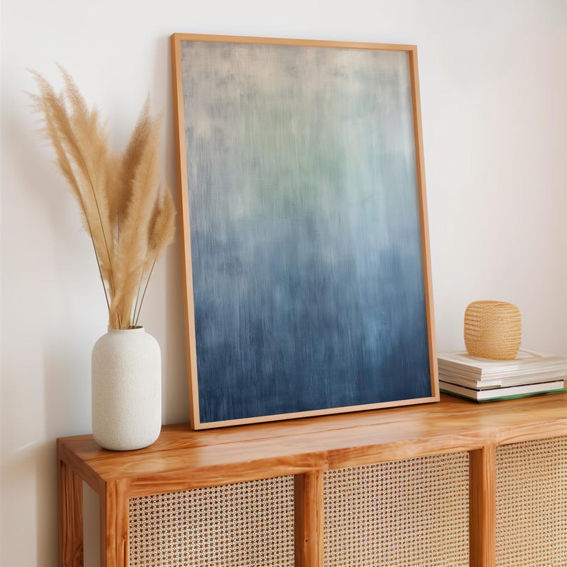 Pode incluir: Uma impress&atilde;o de arte abstrata emoldurada com um gradiente de tons azuis e verdes. A obra de arte &eacute; exibida em um arm&aacute;rio de madeira, ao lado de um vaso branco com capim-dos-pampas seco e uma pilha de livros. A moldura &eacute; de madeira clara.