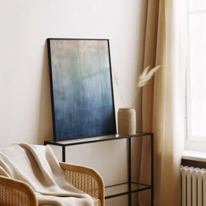 Pode incluir: Uma impress&atilde;o de arte abstrata emoldurada com um gradiente de cores do bege claro ao azul escuro. A obra de arte &eacute; exibida em uma mesa de console de metal preto e vidro. Um vaso bege com plantas secas est&aacute; sobre a mesa. Uma cadeira de vime com um cobertor bege.