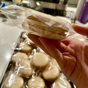 May include: A hand holding a clear plastic bag containing several light-colored macarons. The macarons are round with a creamy filling. A baking sheet with more macarons in individual plastic bags is in the foreground.