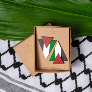 Może przedstawiać: Zestaw drewnianych kolczyków w kształcie trójkątów, pomalowanych kolorami palestyńskiej flagi. Kolczyki są zapakowane w małe kartonowe pudełko z nadrukiem "Umile Wood Things" na spodzie.