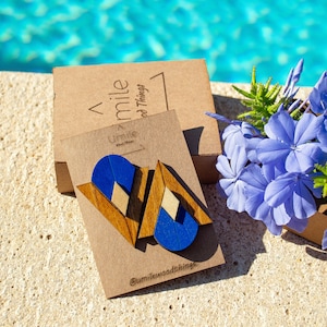Può includere: Un paio di orecchini geometrici in legno con accenti blu e marroni. Gli orecchini sono esposti su una carta marrone con il testo "Umile Wood Things" e "@umilewoodthings".