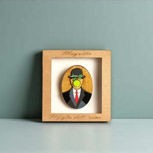 Può includere: Opera d'arte incorniciata in legno raffigurante "Il figlio dell'uomo" di René Magritte. L'opera mostra un uomo in abito con bombetta e una mela verde che gli copre il volto. La cornice riporta il nome dell'artista e il titolo dell'opera.