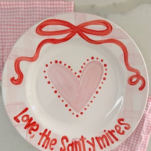 May include: White ceramic plate with a pink heart design, red bow, and the words "Love the Santymires" in red. The plate is decorated with a pink and white gingham pattern and is placed on a pink gingham napkin.