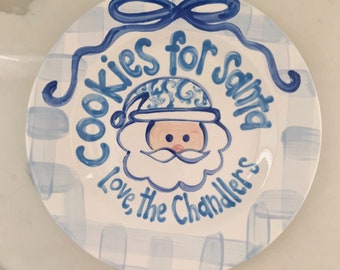 May include: A white ceramic plate with a blue and white checkered pattern. The plate has a blue ribbon and the words "Cookies for Santa Love the Chandlers" written on it. A cartoon Santa Claus face is in the center of the plate.