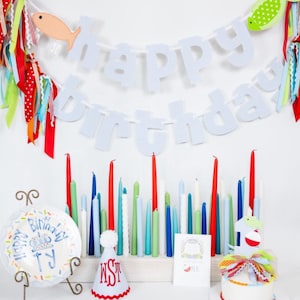 May include: A festive birthday scene with a "Happy Birthday" banner, colorful candles, and a small cake. The banner features blue and white checkered letters and fish-shaped decorations. A plate with birthday text and a party hat are also present.