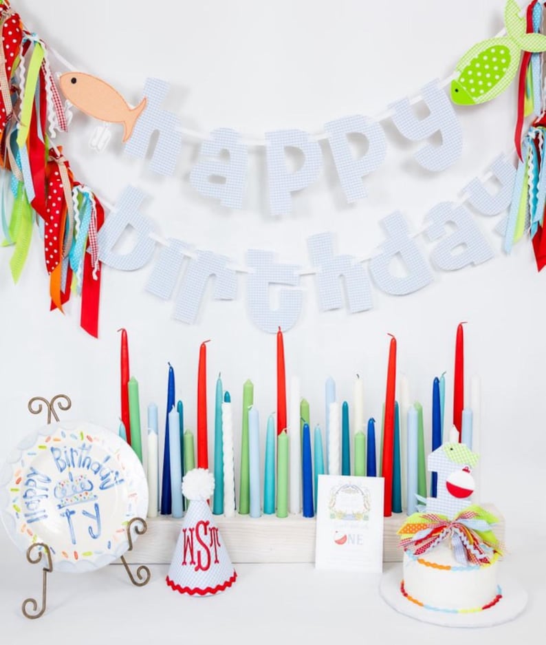 May include: A festive birthday scene with a "Happy Birthday" banner, colorful candles, and a small cake. The banner features blue and white checkered letters and fish-shaped decorations. A plate with birthday text and a party hat are also present.