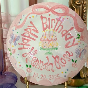May include: A round white ceramic plate with a pink scalloped border, featuring a hand-painted pink cake with candles, butterflies, and colourful dots. The text reads Happy Birthday Hannah Rose and the date 20.10.2025 in pink script lettering.