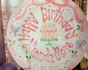 May include: A round white ceramic plate with a pink scalloped border, featuring a hand-painted pink cake with candles, butterflies, and colorful dots. The text reads Happy Birthday Hannah Rose and the date 10.20.2025 in pink script lettering.