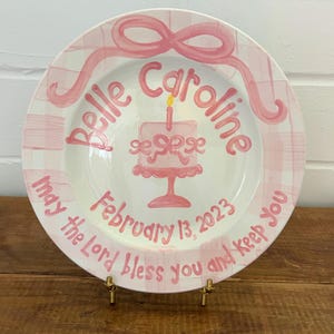 May include: A white ceramic plate with a pink bow and ribbon design. The plate features the name "Belle Caroline", a cake illustration, and the date "February 13, 2023". The text "May the Lord bless you and keep you" is also included.