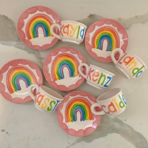 Peut inclure: Ensemble de tasses et soucoupes en céramique blanche. Chaque soucoupe a un bord festonné rose et un motif arc-en-ciel. Les tasses sont personnalisées avec des noms aux couleurs de l'arc-en-ciel. Un service à thé enfantin joyeux et coloré.